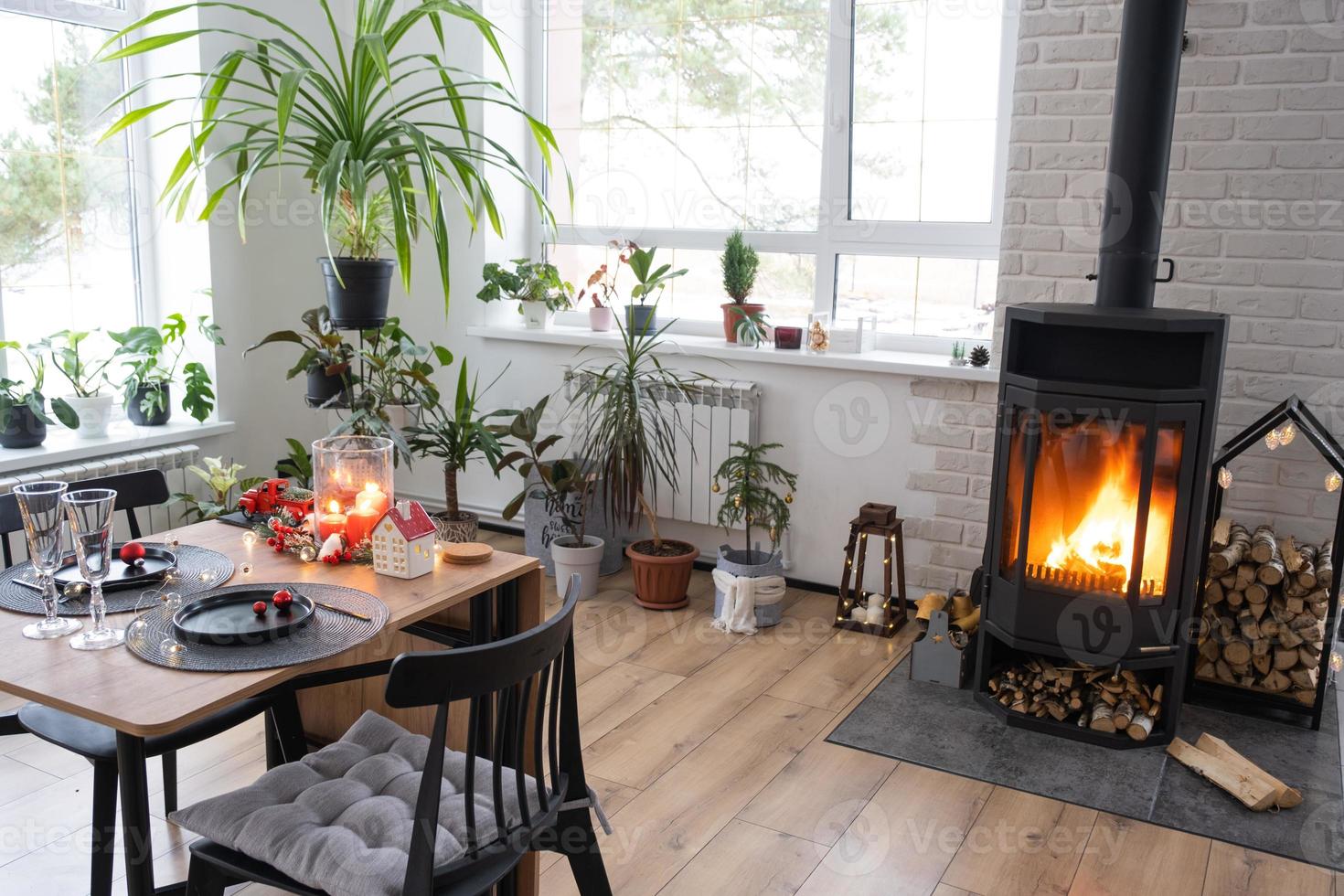 Festive interior of house is decorated for Christmas and New Year in loft style with black stove, fireplace, Christmas tree. Warm studio room with set table, burning wood, cozy and heating of home photo