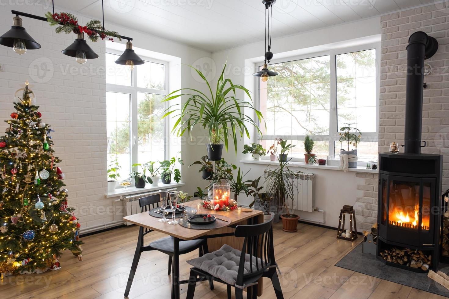 Festive interior of house is decorated for Christmas and New Year in loft style with black stove, fireplace, Christmas tree. Warm studio room with set table, burning wood, cozy and heating of home photo