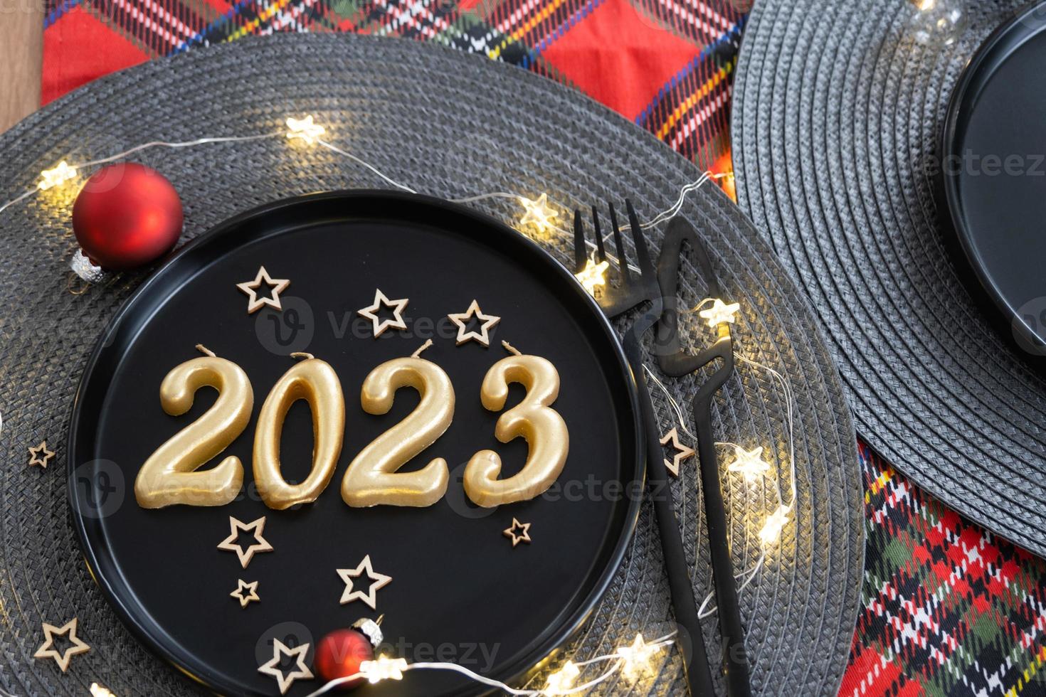 Golden figures 2023 from candles on a black plate on a festive table with a New Year's serving. Loft-style interior, party, feast. Wicker napkin, fork, knife photo