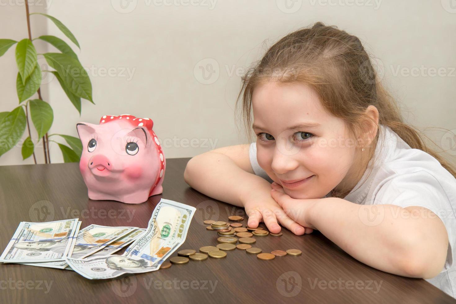 there is a piggy bank on the table, money lies nearby, a smiling baby sits at the table photo