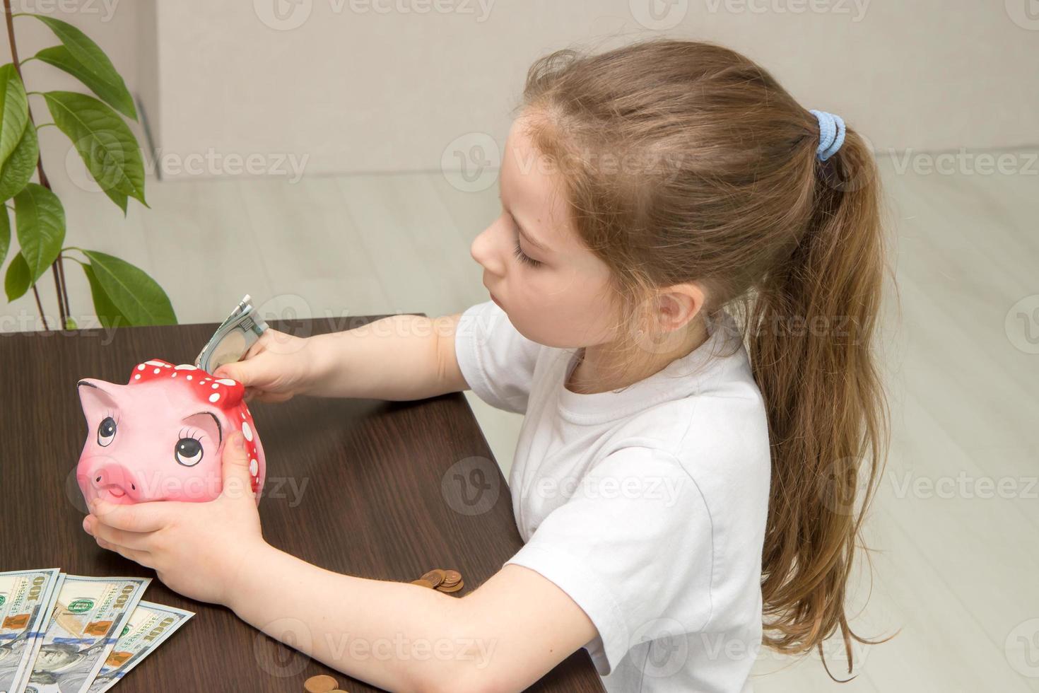 financial literacy concept, cute little girl sitting at the table with concentration puts banknotes into a piggy bank photo