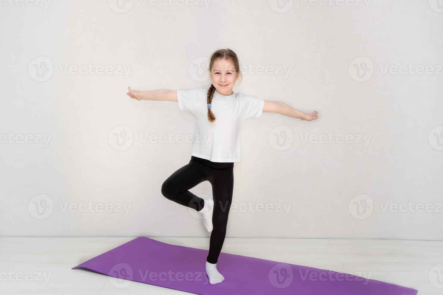 la niña se para en una alfombra de gimnasia en una pierna, tratando de mantener el equilibrio, el fitness de los niños, los deportes foto