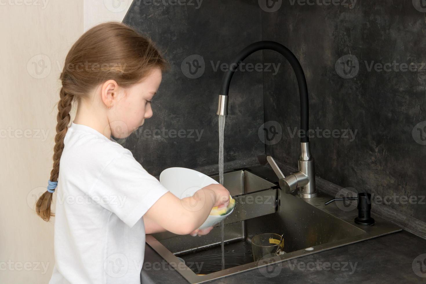 cute little girl with a pigtail washes the dishes in the kitchen at the sink photo