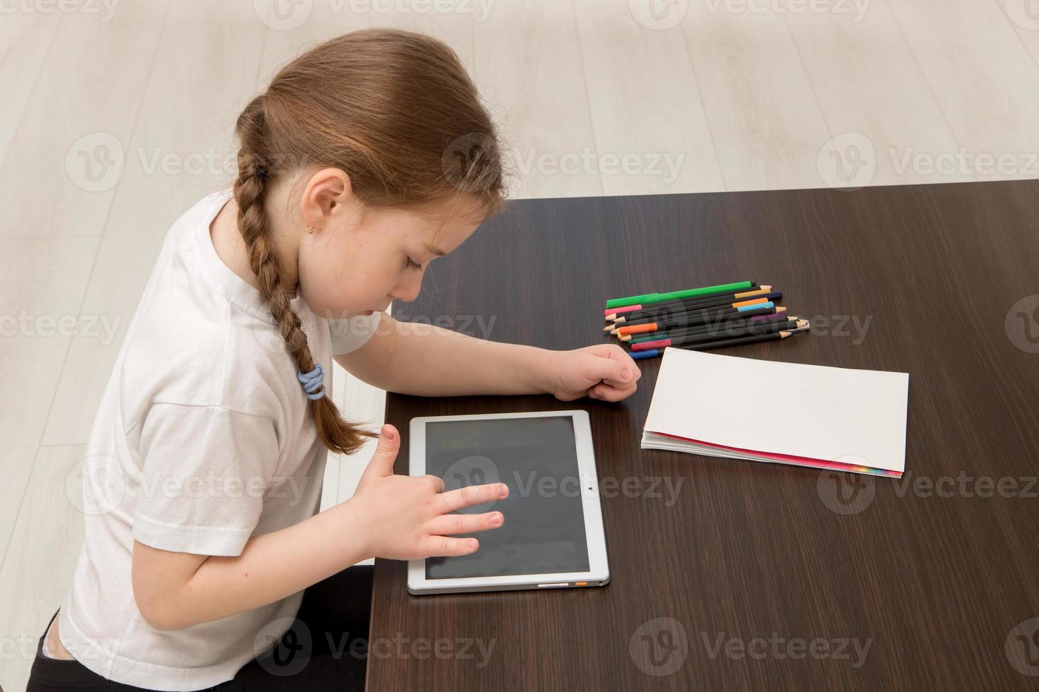 little girl sits at a table and runs her finger across a blank tablet screen next to it lies an empty sheet and colored pencils photo