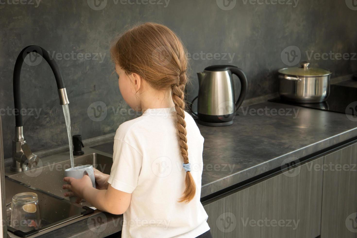 cute little girl with a pigtail washes dishes in the kitchen at the sink photo