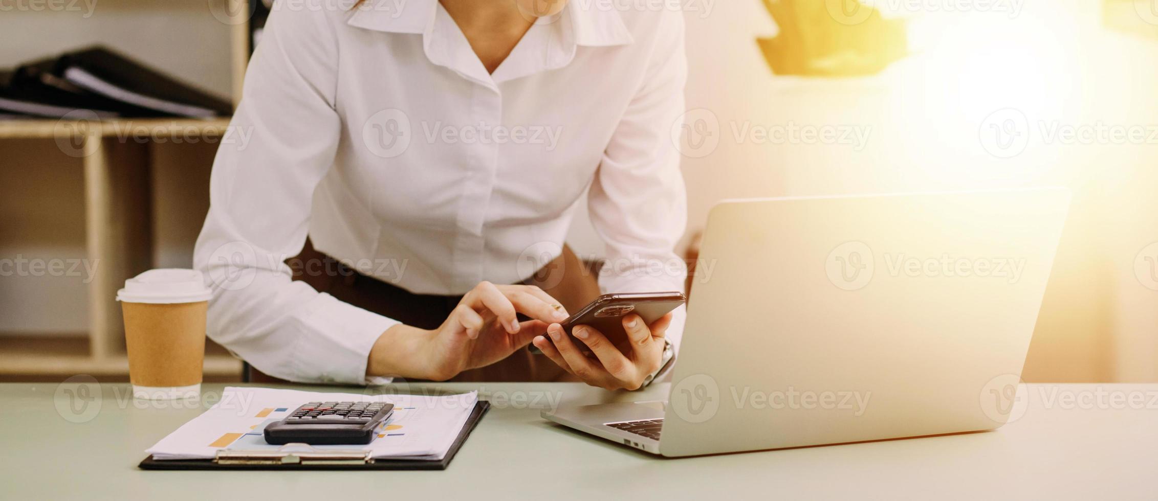 Young business woman sitting in office at table and using smartphone. On desk is laptop and tablet computer, on screen charts and graphs. Woman analyzing data. Student learning online. photo