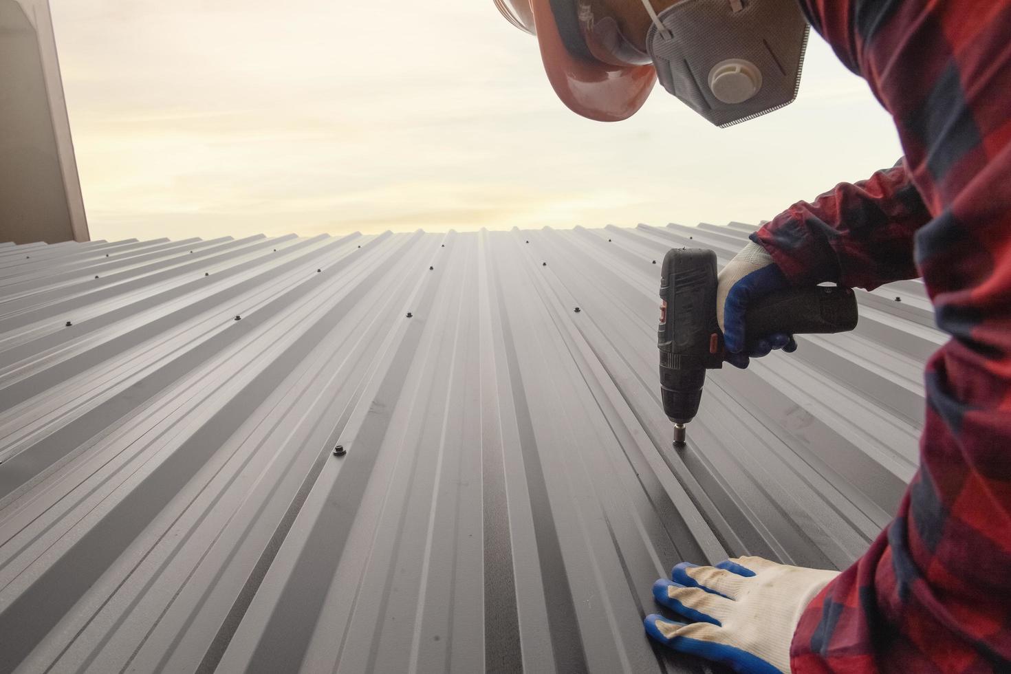 cerrar el trabajador de techado de perforación con uniforme protector y guantes, herramientas de techado, instalación de techos nuevos en construcción, taladro eléctrico usado en techos nuevos con láminas de metal. foto