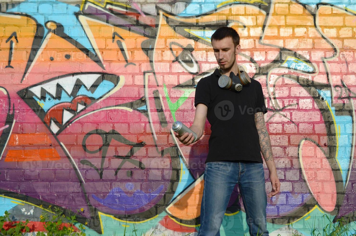 Young caucasian graffiti artist in black t-shirt with silver aerosol spray can near colorful graffiti in pink tones on brick wall. Street art and contemporary painting process photo