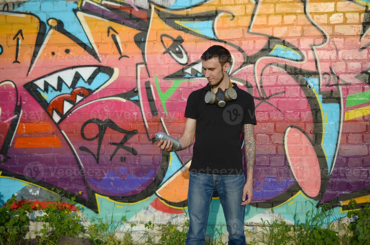 Young caucasian graffiti artist in black t-shirt with silver aerosol spray can near colorful graffiti in pink tones on brick wall. Street art and contemporary painting process photo