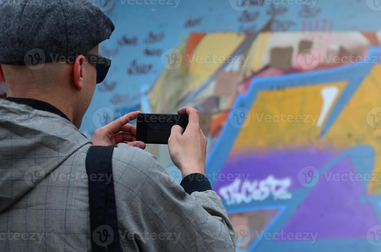 A young graffiti artist photographs his completed picture on the wall. The guy uses modern technology to capture a colorful abstract graffiti drawing. Focus on the photographing device photo