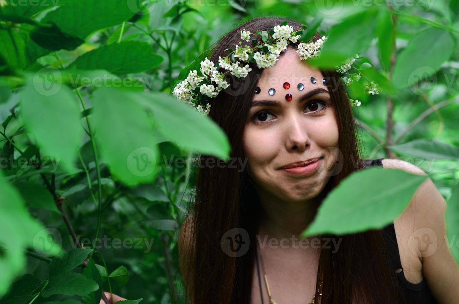 A forest picture of a beautiful young brunette of European appearance with dark brown eyes and large lips. On the girl's head is wearing a floral wreath, on her forehead shiny decorations photo