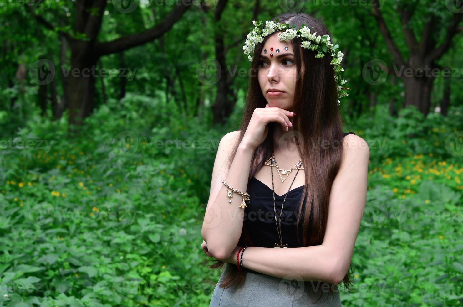 A forest picture of a beautiful young brunette of European appearance with dark brown eyes and large lips. On the girl's head is wearing a floral wreath, on her forehead shiny decorations photo