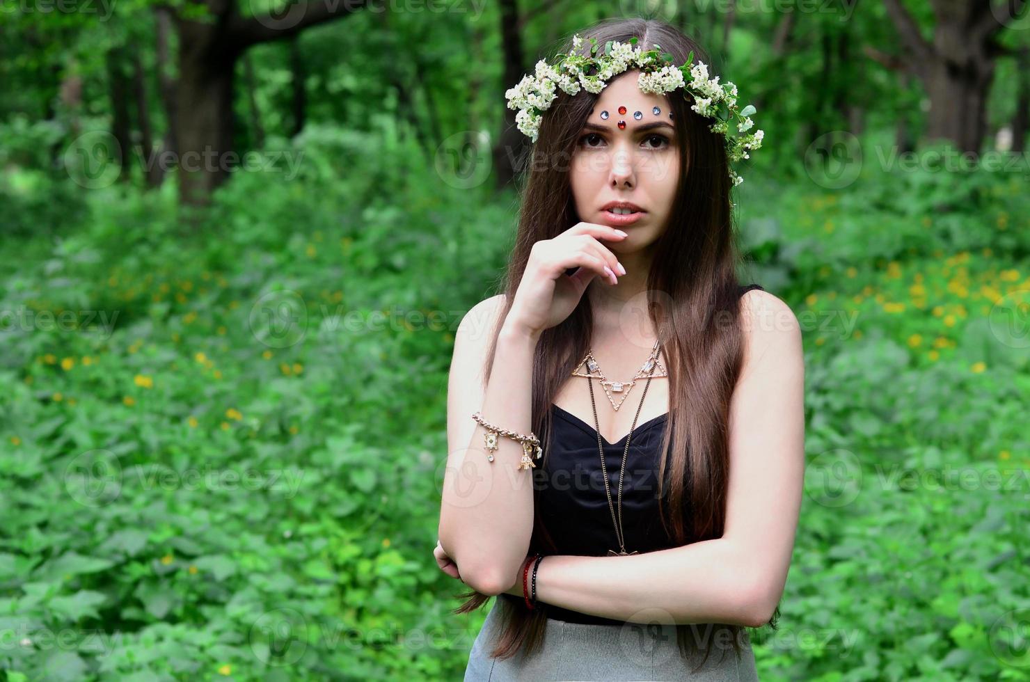Portrait of an emotional young girl with a floral wreath on her head and shiny ornaments on her forehead. Cute brunette posing in a burgeoning beautiful forest in the daytime on a fine day photo