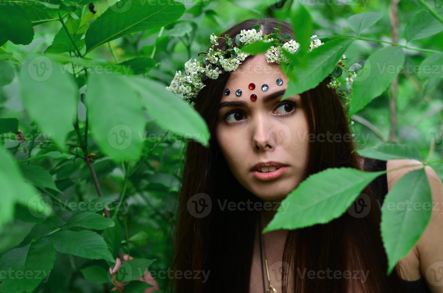 Portrait of an emotional young girl with a floral wreath on her head and shiny ornaments on her forehead. Cute brunette posing in a burgeoning beautiful forest in the daytime on a fine day photo