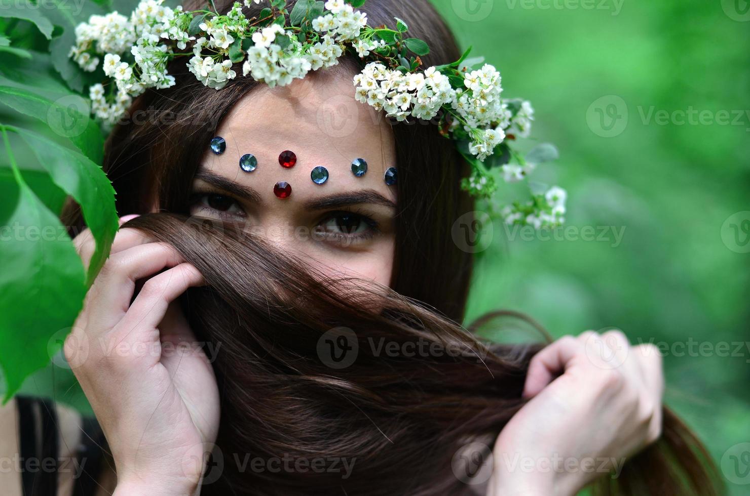 Portrait of an emotional young girl with a floral wreath on her head and shiny ornaments on her forehead. Cute brunette posing in a burgeoning beautiful forest in the daytime on a fine day photo