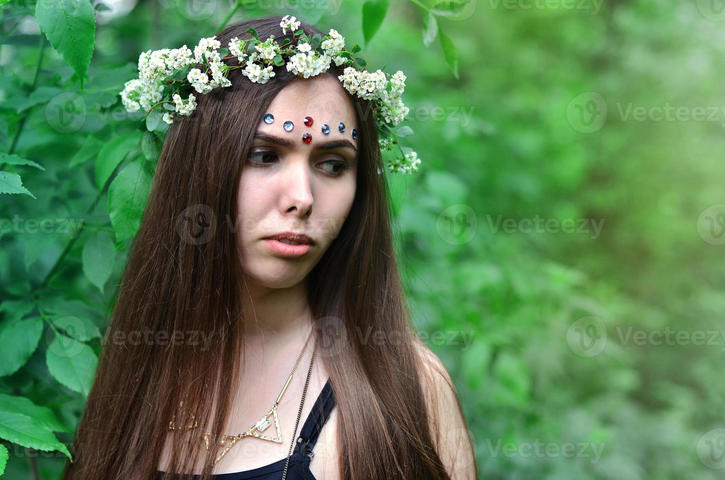 una foto del bosque de una hermosa joven morena de apariencia europea con ojos marrones oscuros y labios grandes. en la cabeza de la niña lleva una corona de flores, en la frente adornos brillantes