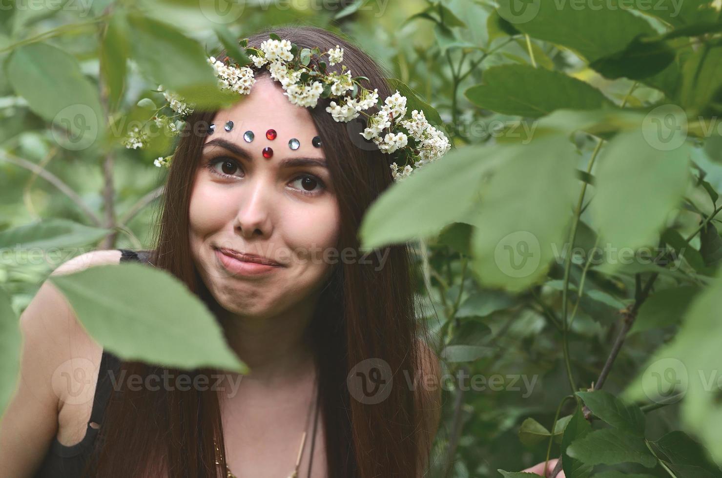 A forest picture of a beautiful young brunette of European appearance with dark brown eyes and large lips. On the girl's head is wearing a floral wreath, on her forehead shiny decorations photo