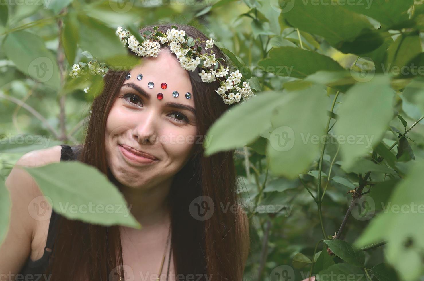 retrato de una joven emocional con una corona floral en la cabeza y adornos brillantes en la frente. linda morena posando en un hermoso bosque floreciente durante el día en un buen día foto