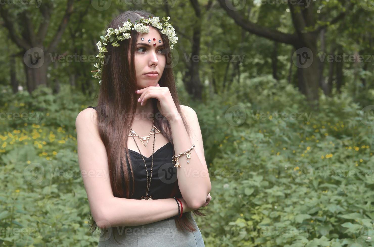 A forest picture of a beautiful young brunette of European appearance with dark brown eyes and large lips. On the girl's head is wearing a floral wreath, on her forehead shiny decorations photo