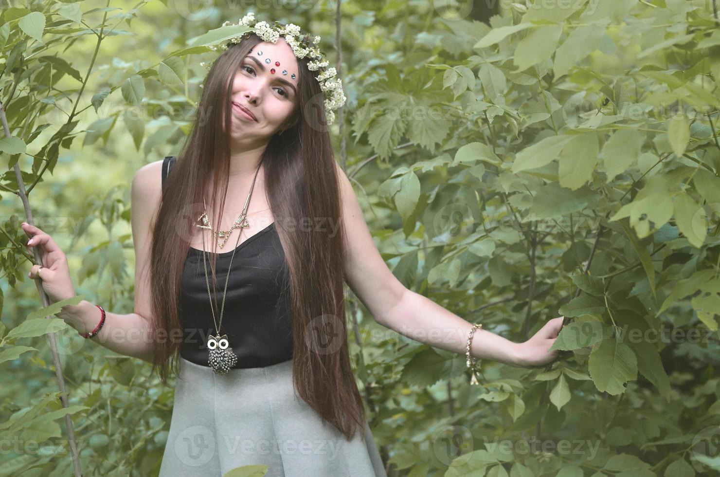 Portrait of an emotional young girl with a floral wreath on her head and shiny ornaments on her forehead. Cute brunette posing in a burgeoning beautiful forest in the daytime on a fine day photo
