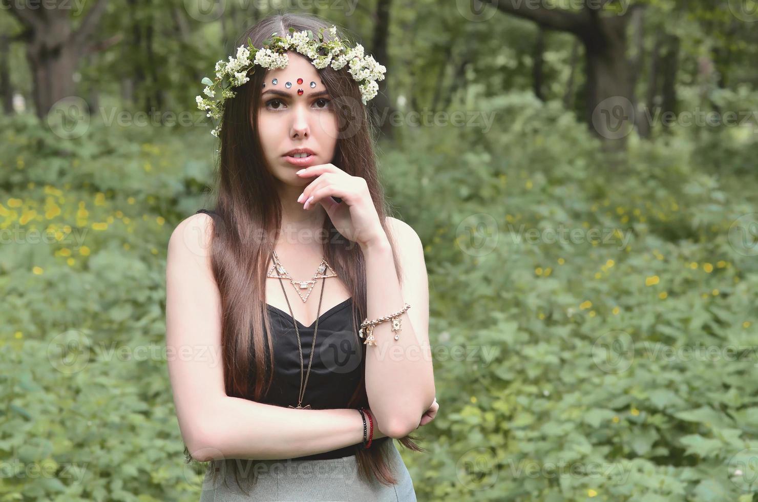 retrato de una joven emocional con una corona floral en la cabeza y adornos brillantes en la frente. linda morena posando en un hermoso bosque floreciente durante el día en un buen día foto