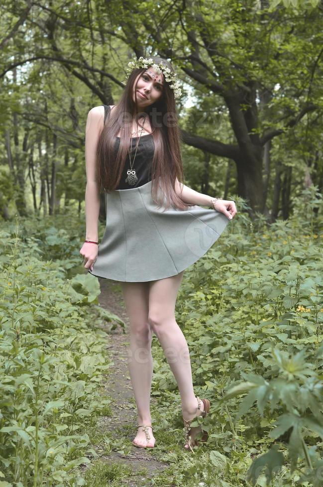 Portrait of an emotional young girl with a floral wreath on her head and shiny ornaments on her forehead. Cute brunette posing in a burgeoning beautiful forest in the daytime on a fine day photo