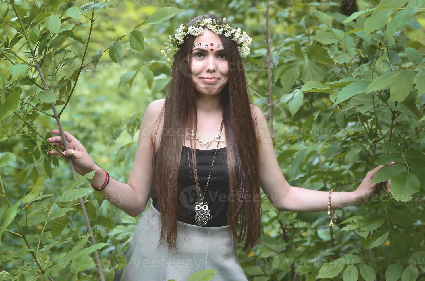 Portrait of an emotional young girl with a floral wreath on her head and shiny ornaments on her forehead. Cute brunette posing in a burgeoning beautiful forest in the daytime on a fine day photo