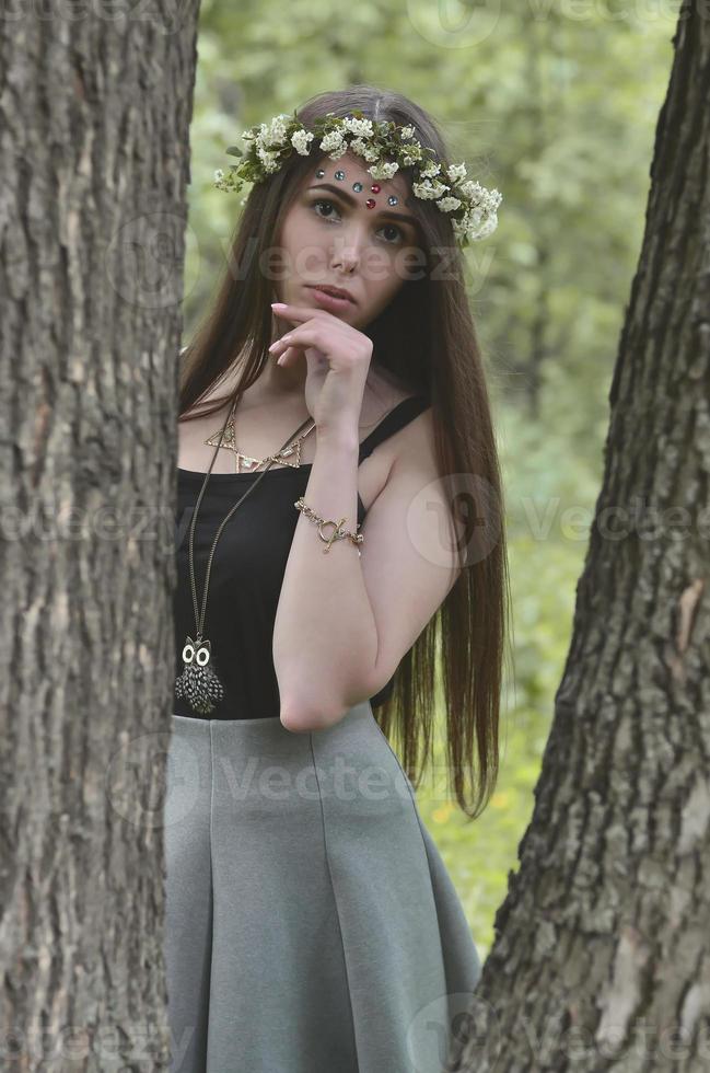 una foto del bosque de una hermosa joven morena de apariencia europea con ojos marrones oscuros y labios grandes. en la cabeza de la niña lleva una corona de flores, en la frente adornos brillantes