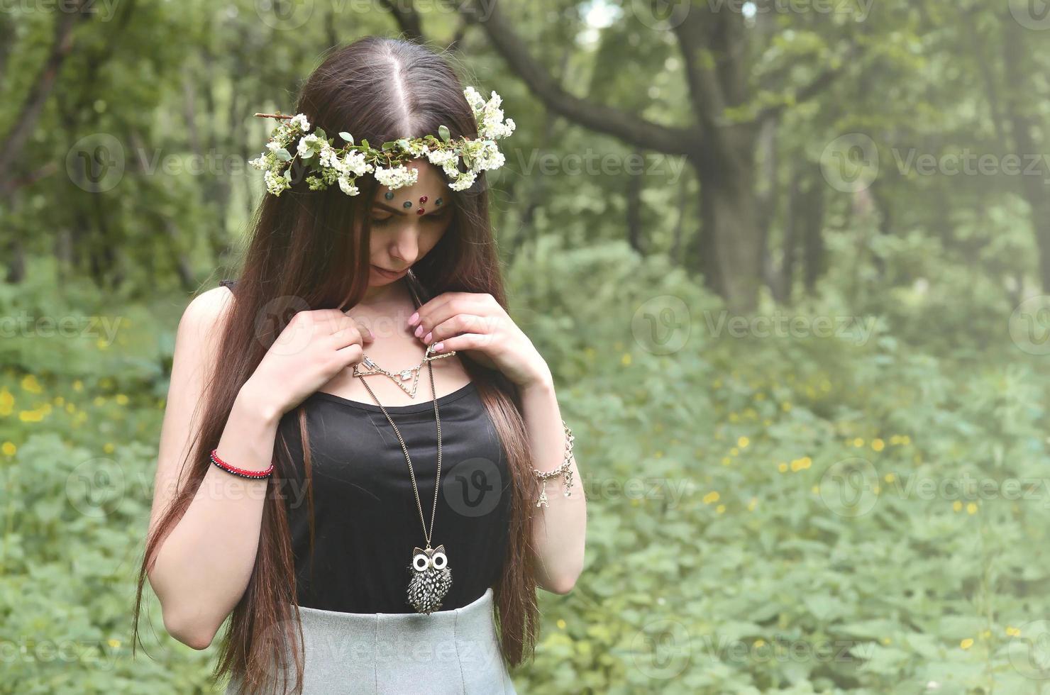 una foto del bosque de una hermosa joven morena de apariencia europea con ojos marrones oscuros y labios grandes. en la cabeza de la niña lleva una corona de flores, en la frente adornos brillantes