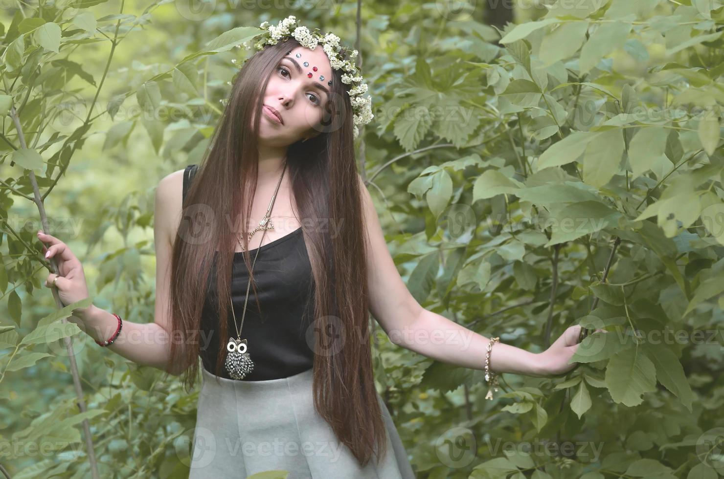 A forest picture of a beautiful young brunette of European appearance with dark brown eyes and large lips. On the girl's head is wearing a floral wreath, on her forehead shiny decorations photo