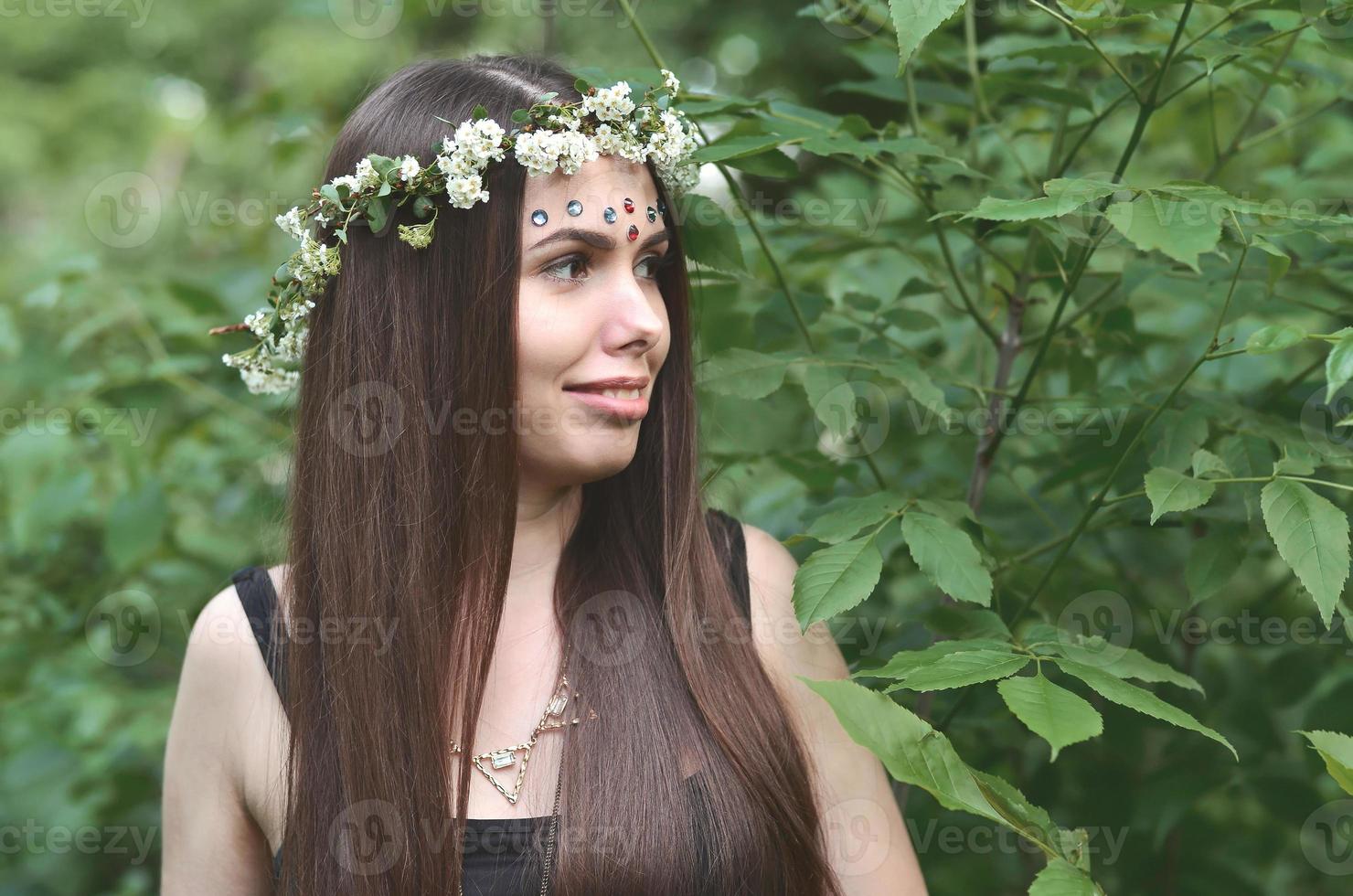 una foto del bosque de una hermosa joven morena de apariencia europea con ojos marrones oscuros y labios grandes. en la cabeza de la niña lleva una corona de flores, en la frente adornos brillantes