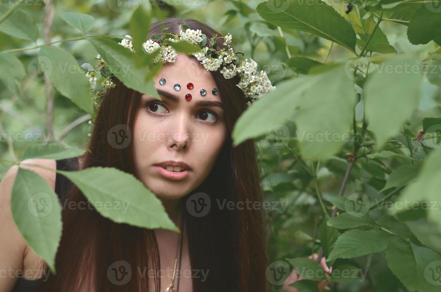 Portrait of an emotional young girl with a floral wreath on her head and shiny ornaments on her forehead. Cute brunette posing in a burgeoning beautiful forest in the daytime on a fine day photo