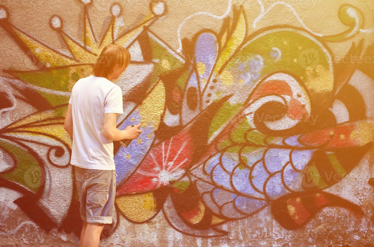 Photo of a young guy in denim shorts and a white shirt. The guy draws on the graffiti wall a drawing with aerosol paints of various colors. The concept of hooliganism and damage to property