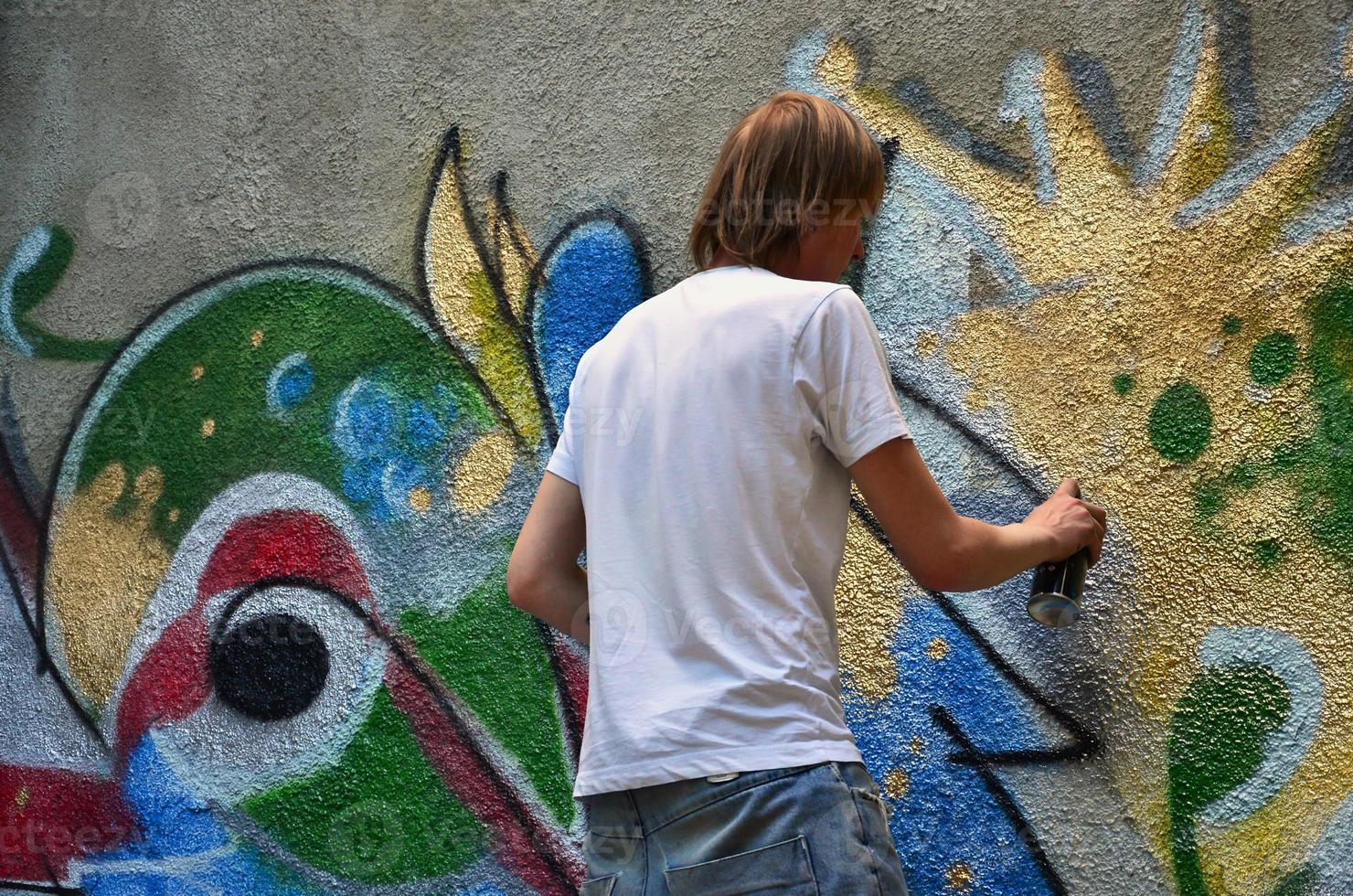 Photo in the process of drawing a graffiti pattern on an old concrete wall. Young long-haired blond guy draws an abstract drawing of different colors. Street art and vandalism concept