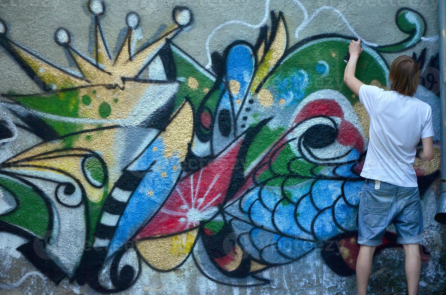 Photo of a young guy in denim shorts and a white shirt. The guy draws on the graffiti wall a drawing with aerosol paints of various colors. The concept of hooliganism and damage to property