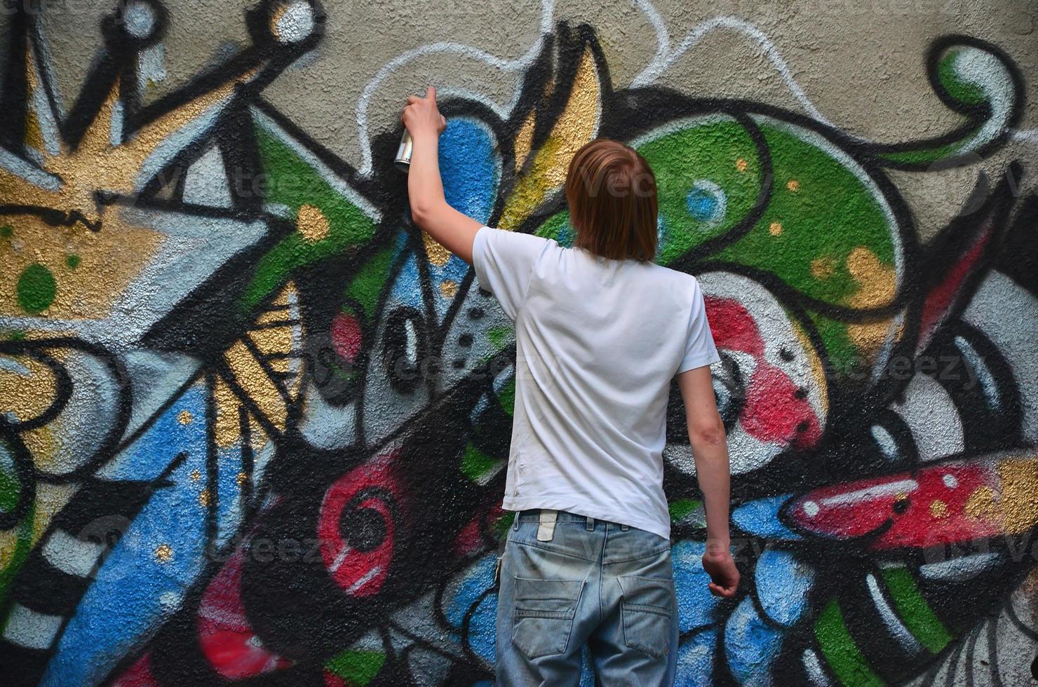 Photo in the process of drawing a graffiti pattern on an old concrete wall. Young long-haired blond guy draws an abstract drawing of different colors. Street art and vandalism concept