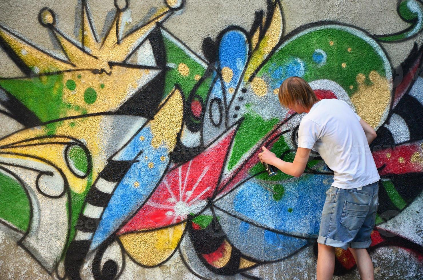 Photo in the process of drawing a graffiti pattern on an old concrete wall. Young long-haired blond guy draws an abstract drawing of different colors. Street art and vandalism concept