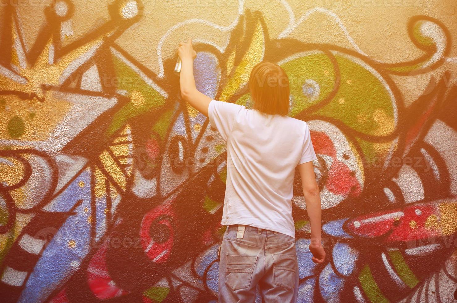 Photo in the process of drawing a graffiti pattern on an old concrete wall. Young long-haired blond guy draws an abstract drawing of different colors. Street art and vandalism concept