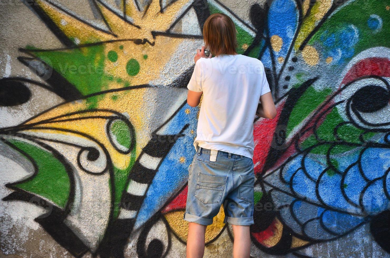 Photo in the process of drawing a graffiti pattern on an old concrete wall. Young long-haired blond guy draws an abstract drawing of different colors. Street art and vandalism concept