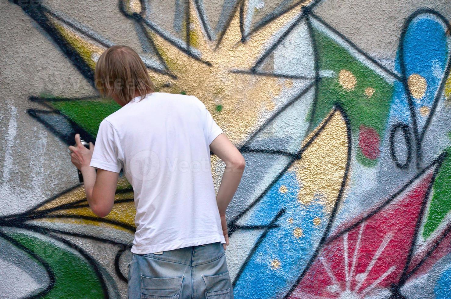 foto en el proceso de dibujar un patrón de graffiti en un antiguo muro de hormigón. un joven rubio de pelo largo dibuja un dibujo abstracto de diferentes colores. concepto de arte callejero y vandalismo