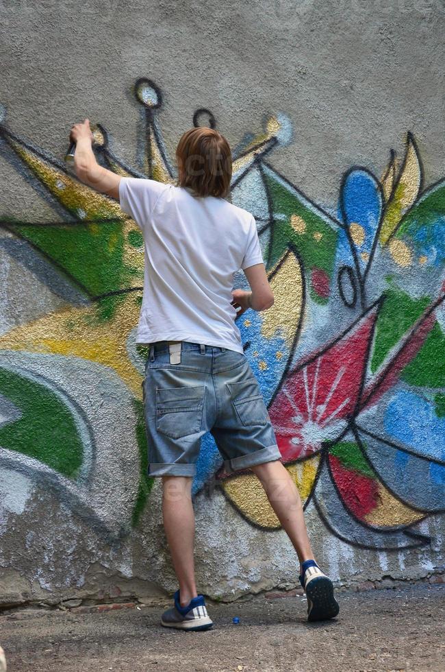 Photo of a young guy in denim shorts and a white shirt. The guy draws on the graffiti wall a drawing with aerosol paints of various colors. The concept of hooliganism and damage to property