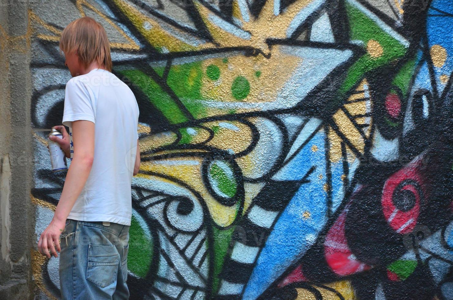 Photo in the process of drawing a graffiti pattern on an old concrete wall. Young long-haired blond guy draws an abstract drawing of different colors. Street art and vandalism concept
