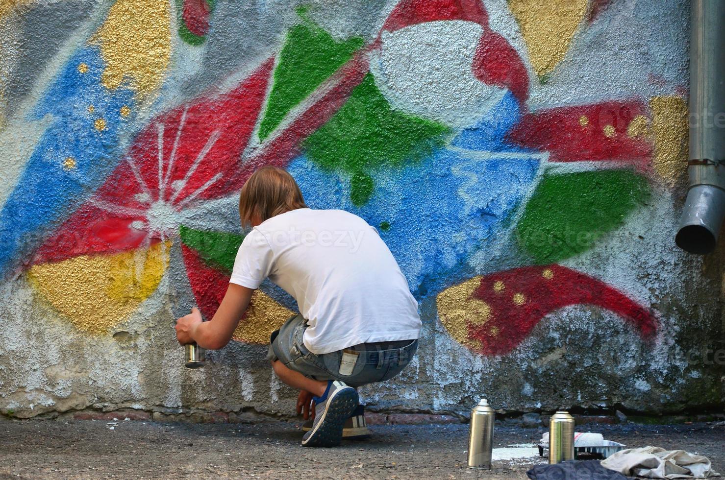 Photo in the process of drawing a graffiti pattern on an old concrete wall. Young long-haired blond guy draws an abstract drawing of different colors. Street art and vandalism concept