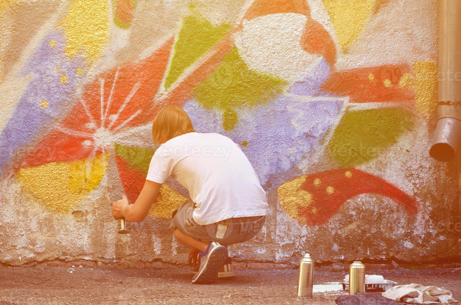 Photo in the process of drawing a graffiti pattern on an old concrete wall. Young long-haired blond guy draws an abstract drawing of different colors. Street art and vandalism concept