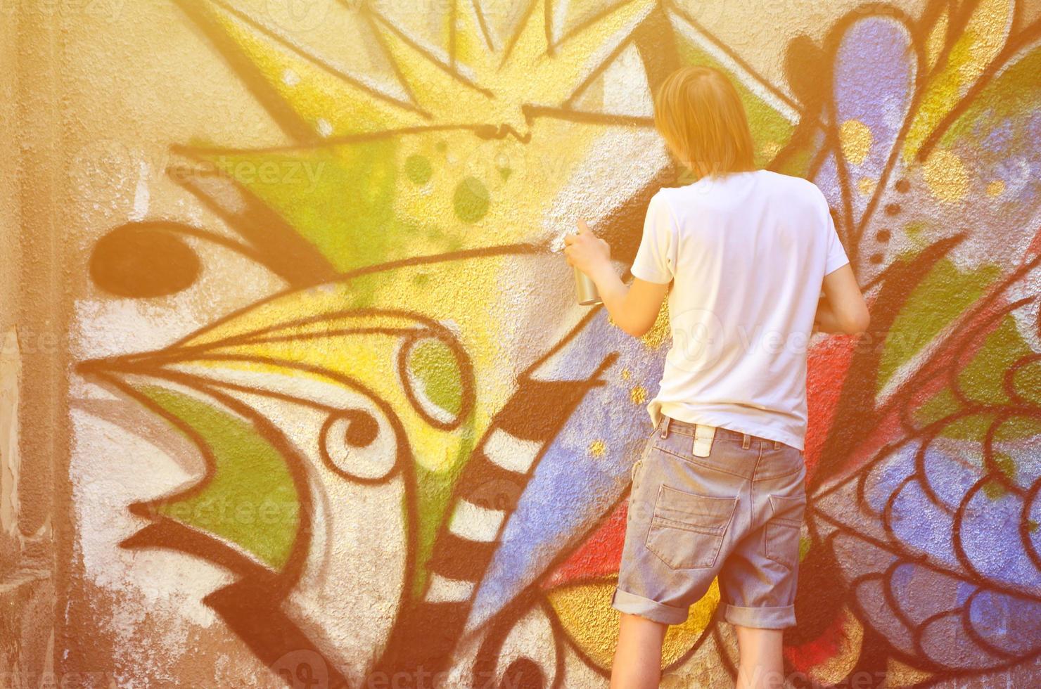 Photo of a young guy in denim shorts and a white shirt. The guy draws on the graffiti wall a drawing with aerosol paints of various colors. The concept of hooliganism and damage to property