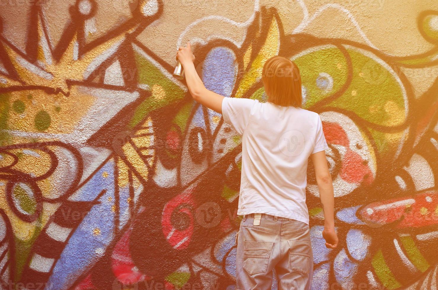 Photo of a young guy in denim shorts and a white shirt. The guy draws on the graffiti wall a drawing with aerosol paints of various colors. The concept of hooliganism and damage to property