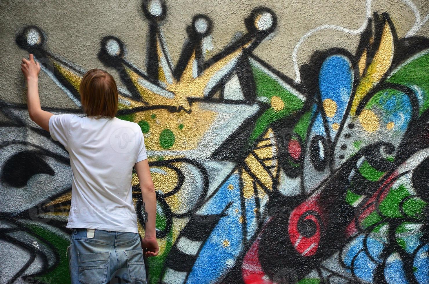 Photo of a young guy in denim shorts and a white shirt. The guy draws on the graffiti wall a drawing with aerosol paints of various colors. The concept of hooliganism and damage to property