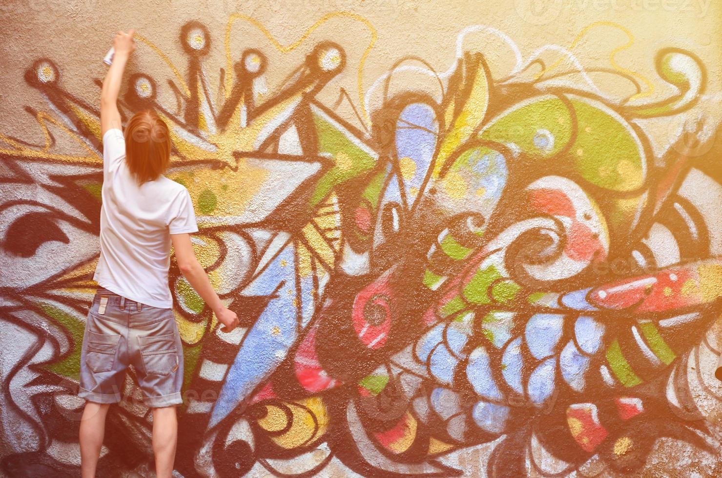 Photo in the process of drawing a graffiti pattern on an old concrete wall. Young long-haired blond guy draws an abstract drawing of different colors. Street art and vandalism concept