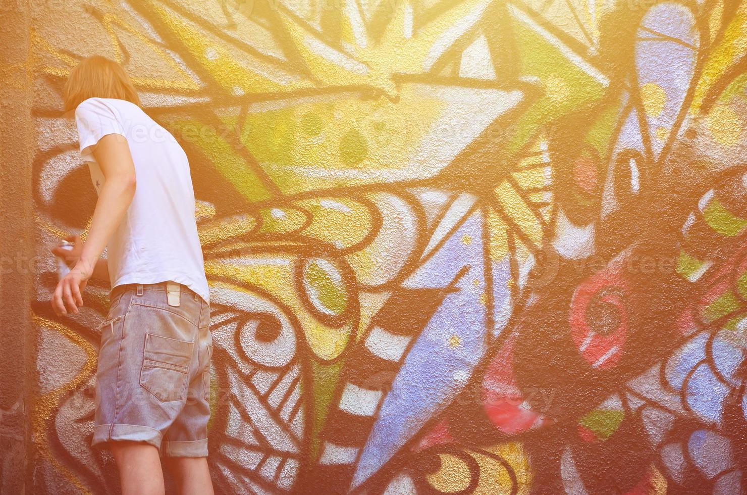 Photo in the process of drawing a graffiti pattern on an old concrete wall. Young long-haired blond guy draws an abstract drawing of different colors. Street art and vandalism concept