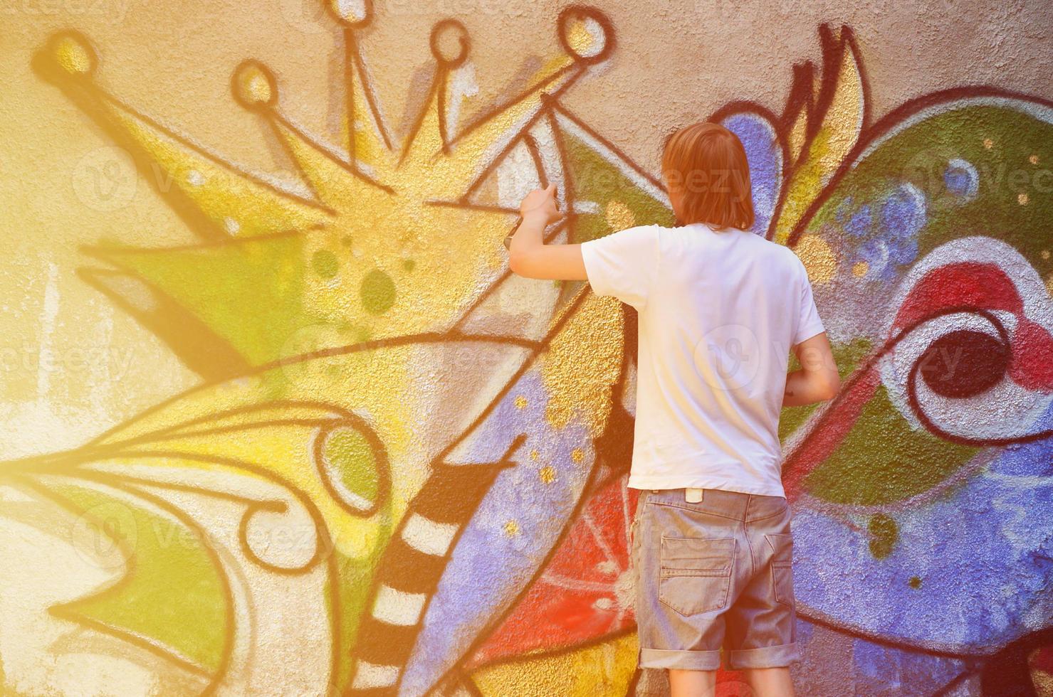 Photo of a young guy in denim shorts and a white shirt. The guy draws on the graffiti wall a drawing with aerosol paints of various colors. The concept of hooliganism and damage to property